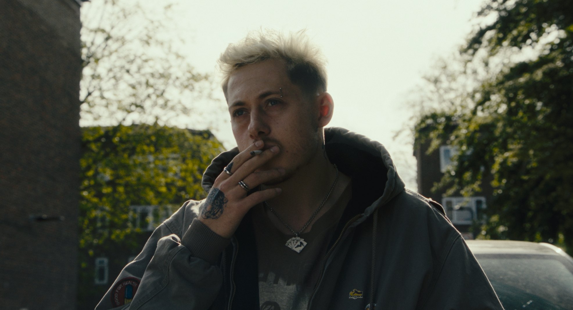 A man with bleached hair smokes a cigarette outdoors in daylight, hand to his mouth.