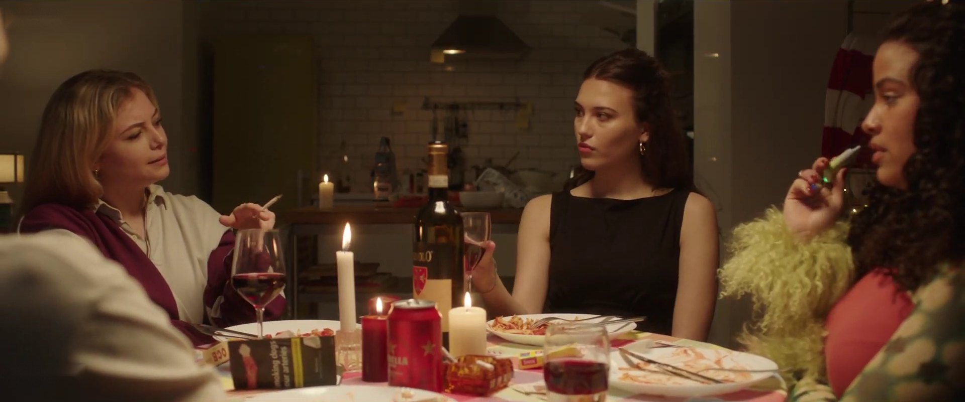 Three women sit around a candlelit dinner table with wine and cigarettes, one looking across at another.