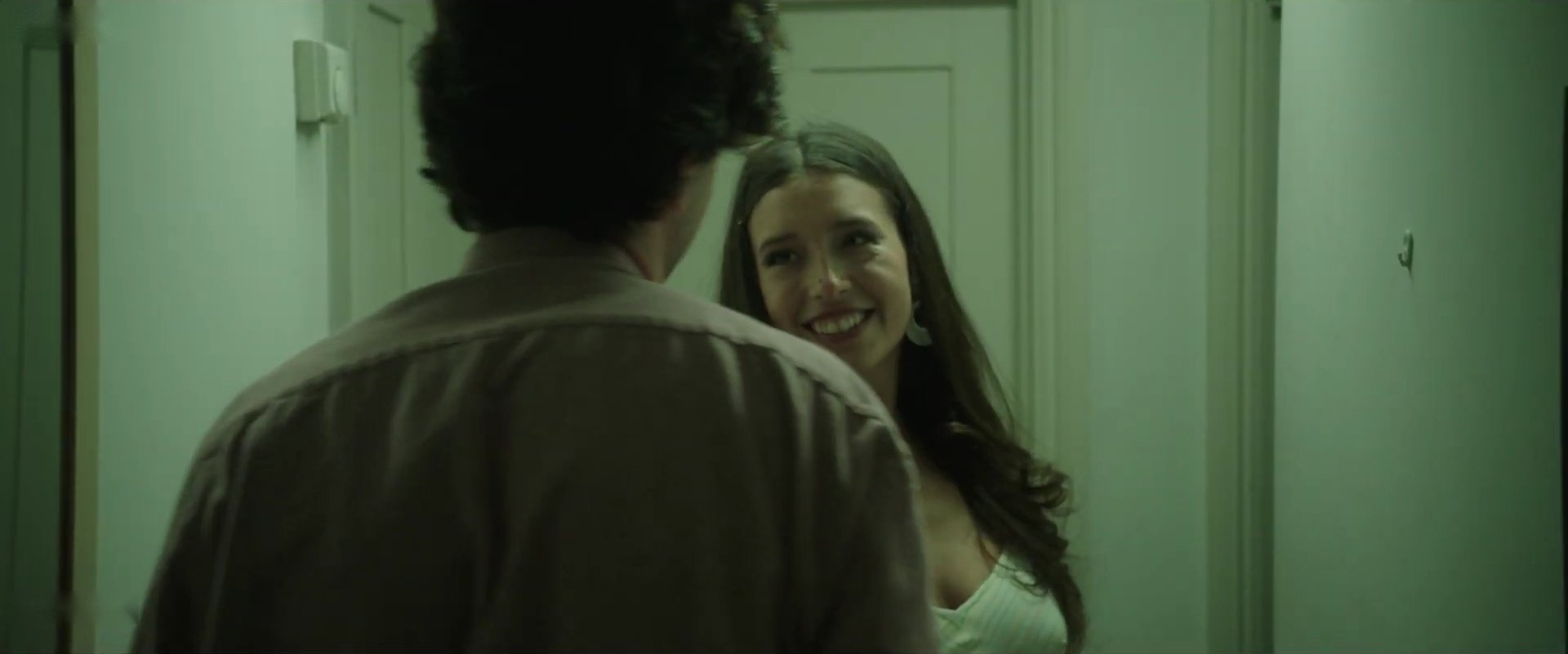A woman with long hair smiles at a man seen from behind, in a green-lit hallway.