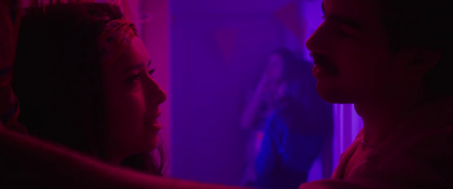 Two faces close together in magenta and blue light, a woman at left and a man with a moustache at right.