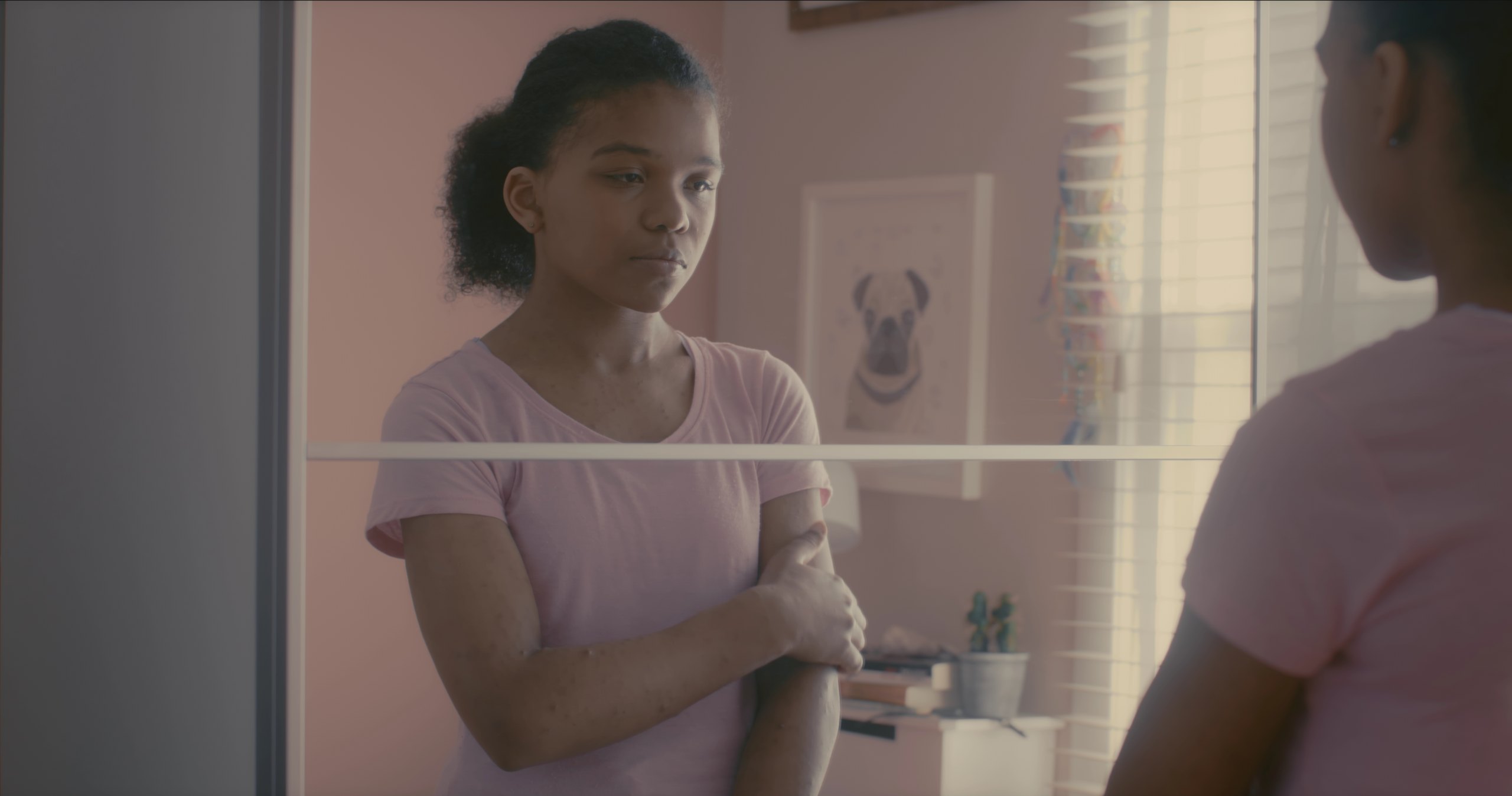 A teenage girl in a pink T-shirt stands with her arms crossed, looking at her reflection in a mirrored wardrobe in a pink bedroom.