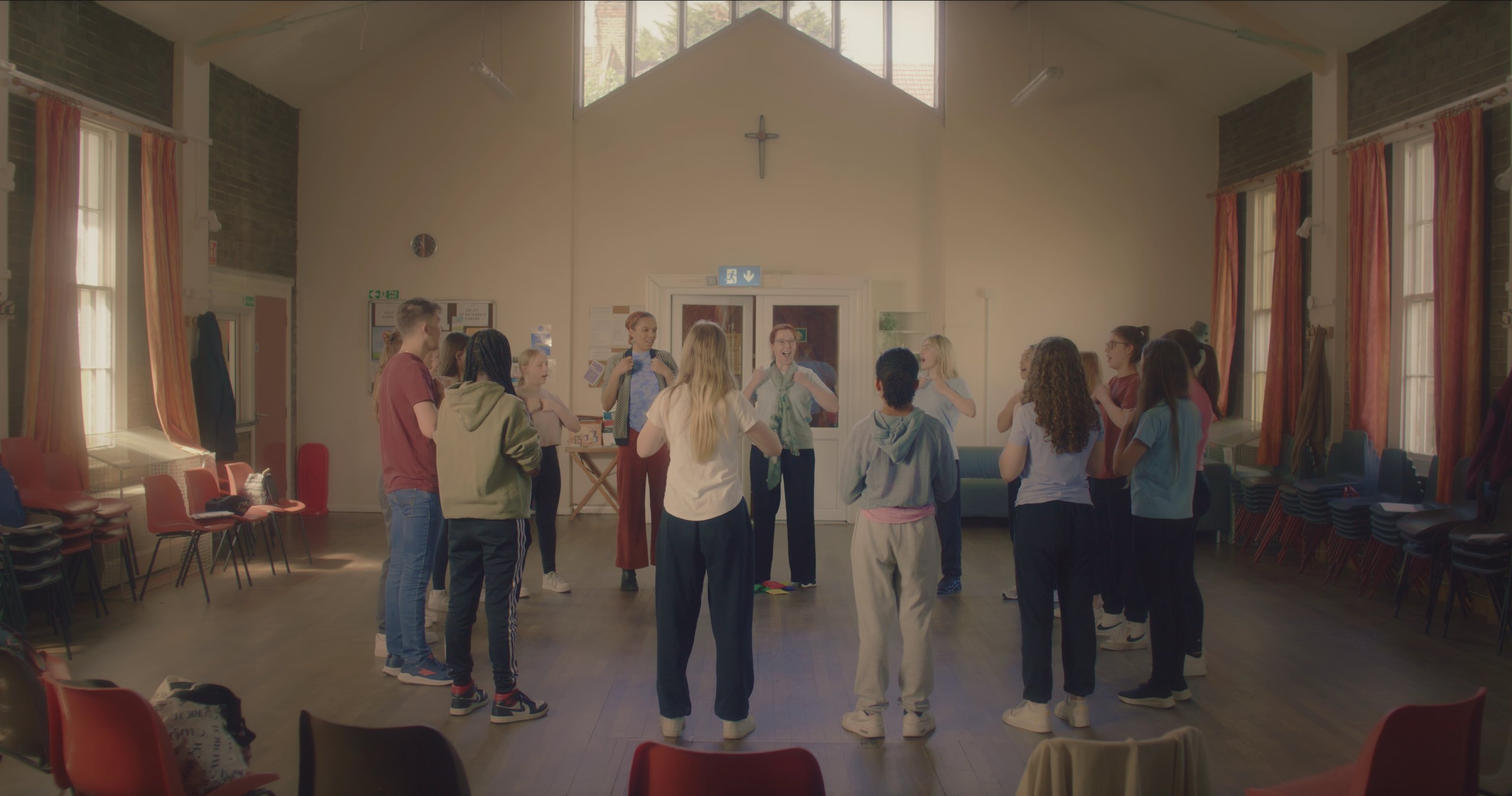 A group of young people and two adults stand in a circle in a community hall, with hands to their chests.