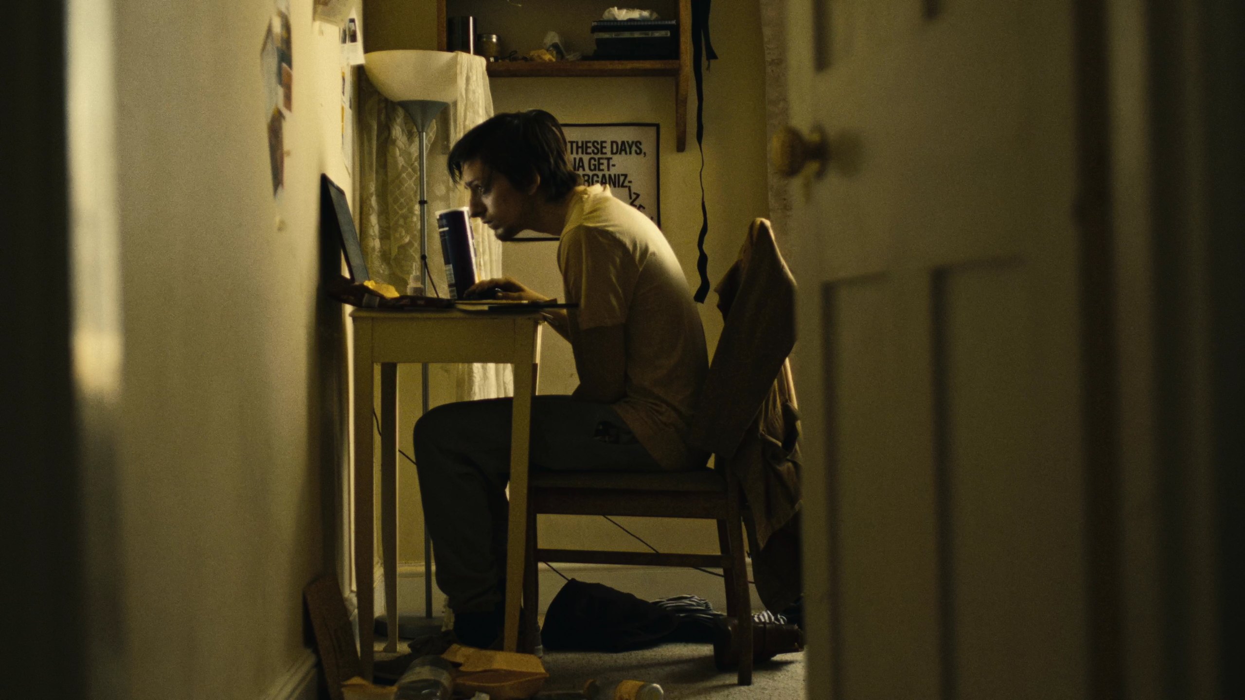 A young man hunched over a laptop at a small desk in a narrow, dimly lit room, seen through a doorway.