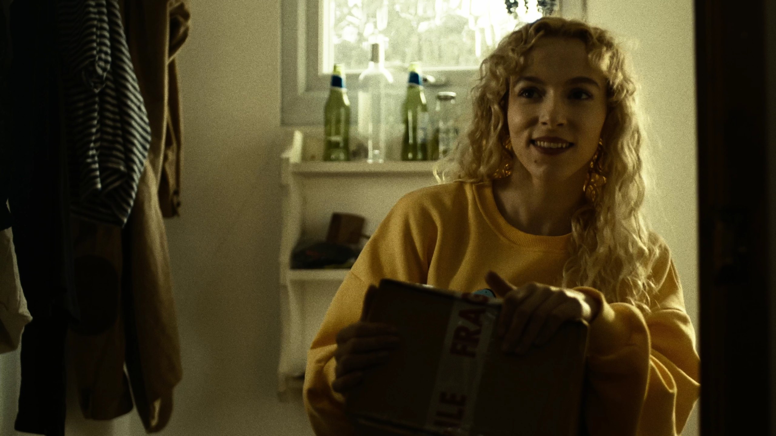 A woman with blonde curly hair in a yellow sweatshirt smiles while holding a cardboard box marked fragile.
