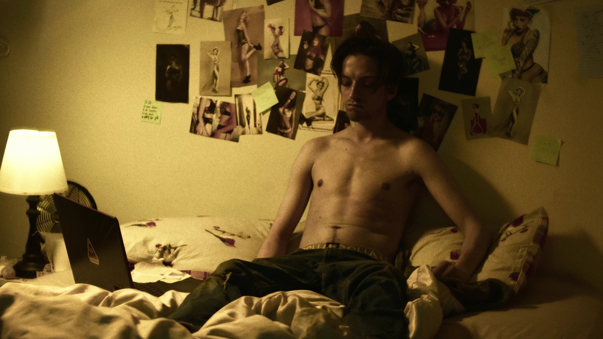 A shirtless man sits against a bedroom wall covered in photographs, with a laptop beside him.
