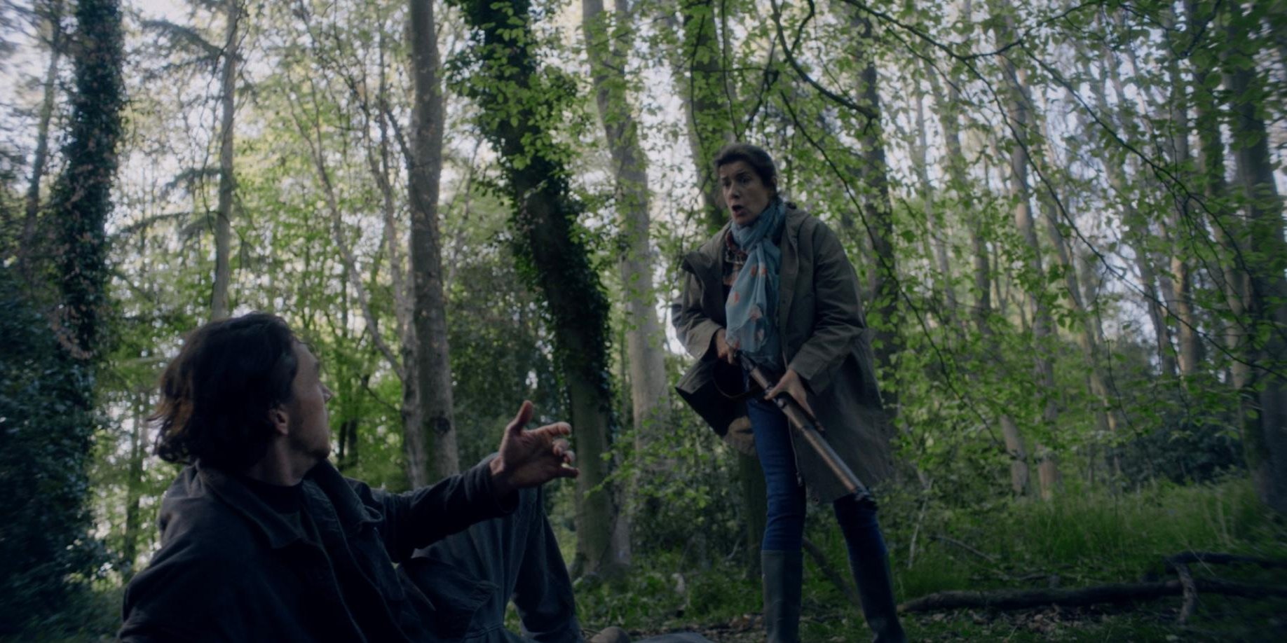 A woman in a green scarf and coat stands holding a shotgun in a sunlit wood, while a man sits on the ground reaching a hand toward her.