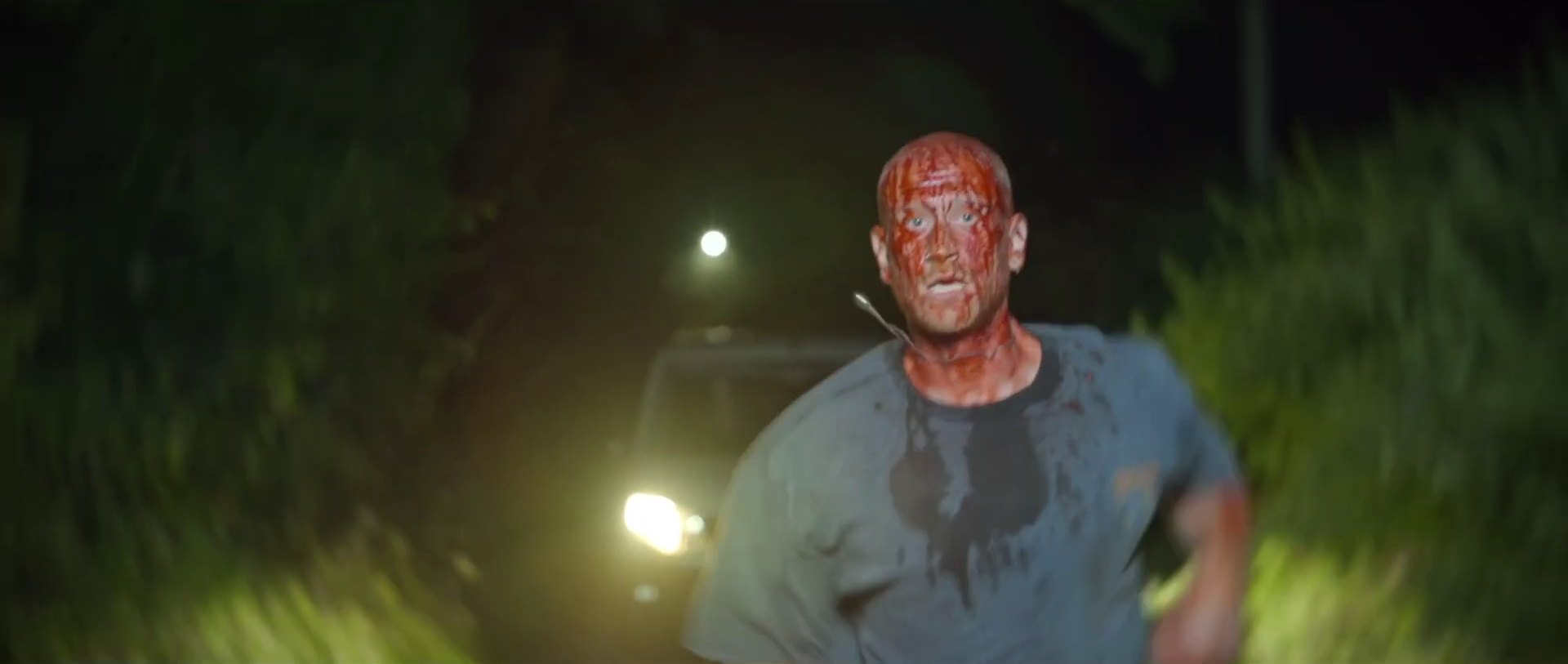 A bald man with blood across his face and shirt runs toward the camera down a dark country lane, headlights behind him.