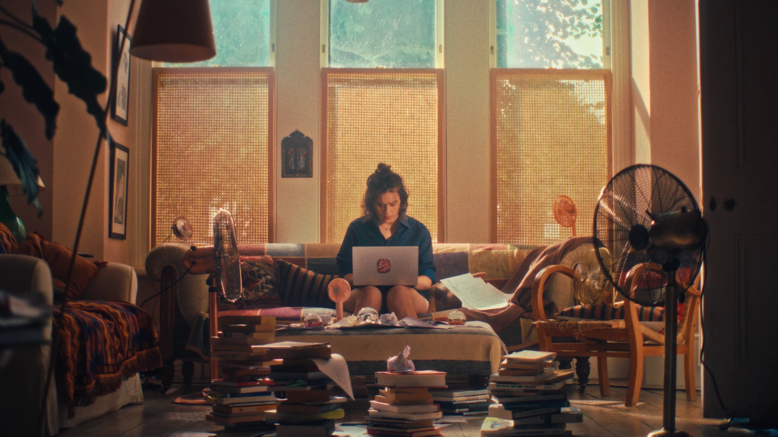 A woman with a topknot works on a laptop on a sofa in a sunlit living room, surrounded by tall stacks of books and electric fans.