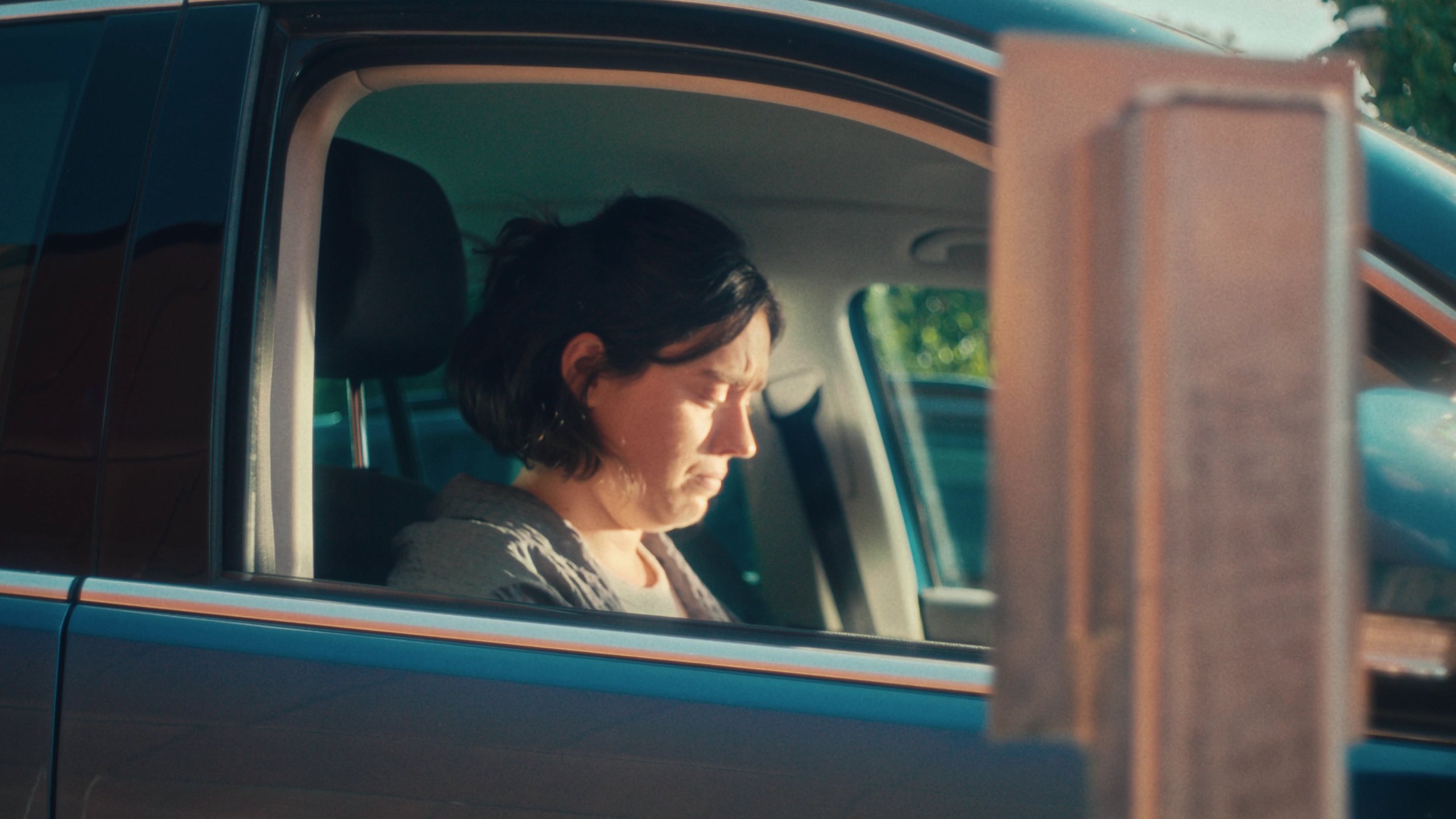 A woman with dark hair sits in a car and cries, seen through the open window in warm sunlight.