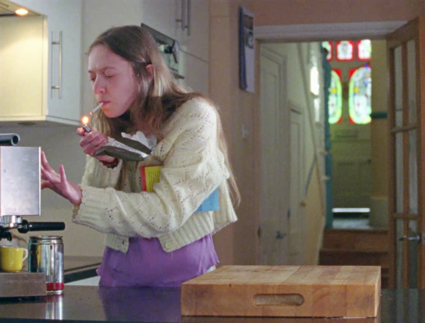 A young woman in a cream cardigan lights a cigarette in a kitchen while holding a book and a packet under her arm.