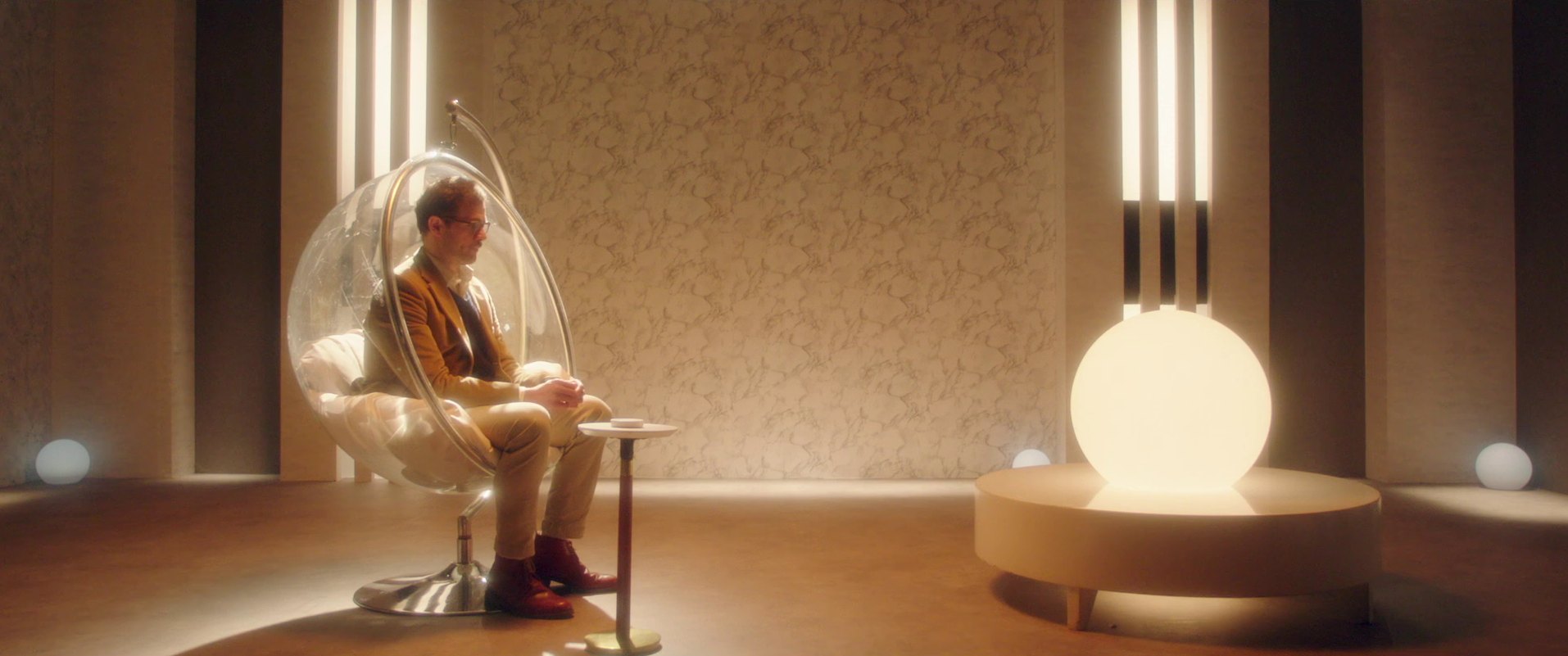 A man in glasses and a mustard jacket sits in a clear bubble chair in a warm, minimal room beside a large glowing globe lamp.